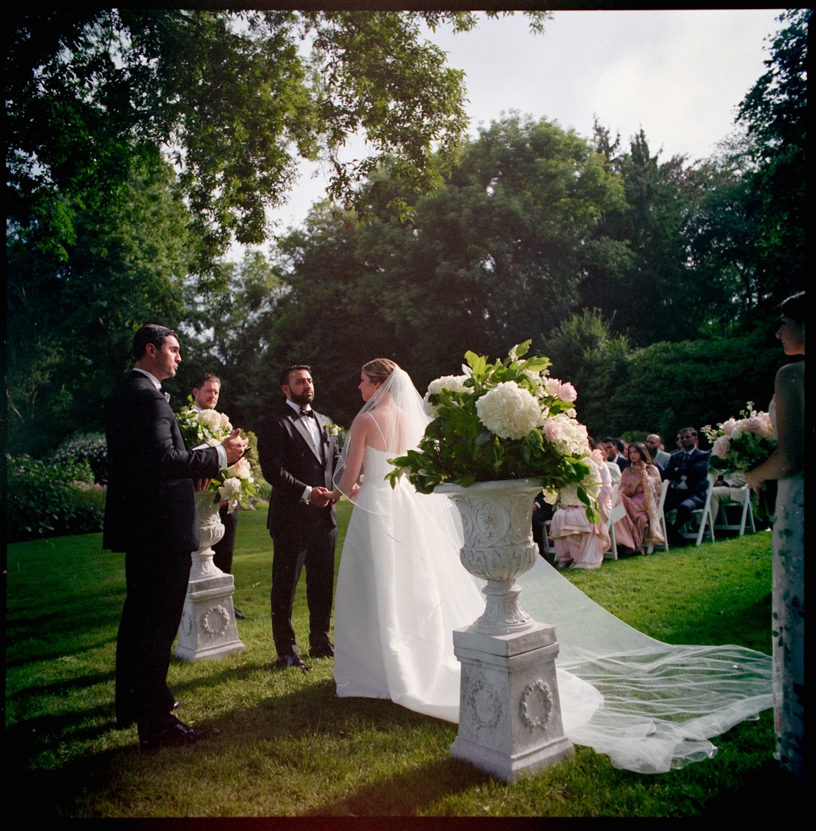 A bride and groom stand holding hands during an outdoor Glen Manor House Wedding Photography ceremony, facing an officiant. Guests are seated in the background, with large flower arrangements decorating the grassy setting.
