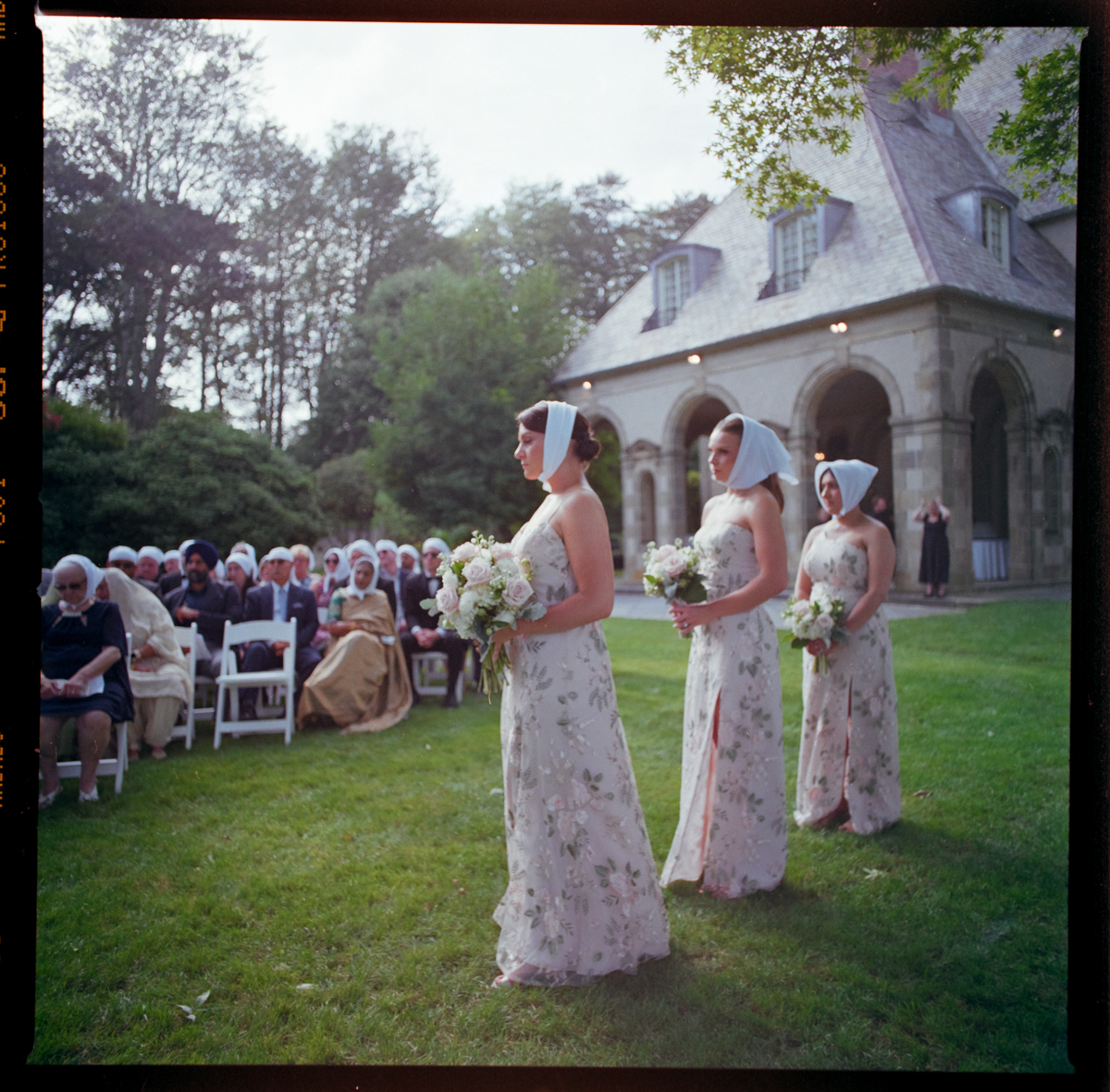 Three bridesmaids in floral dresses with white headscarves stand outdoors holding bouquets, facing a seated crowd at a Glen Manor House Wedding Photography session near a stone building with arches and a sloped roof.