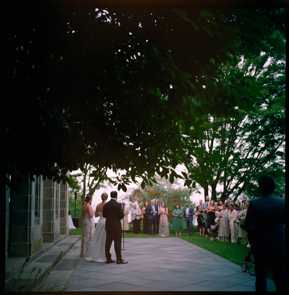 A Glen Manor House Wedding Photography scene captures a couple standing together near the building, addressing guests on a patio nestled among trees and lush greenery during their outdoor ceremony.