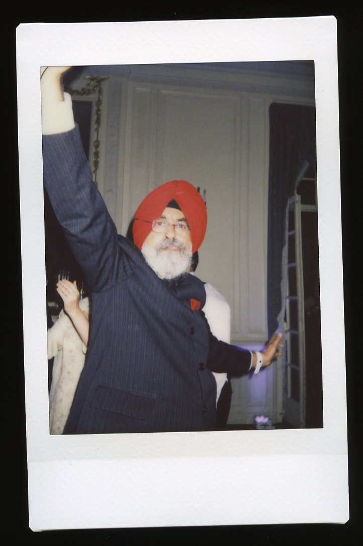 A man wearing a red turban and glasses is smiling and raising one arm while dancing at an indoor event. Captured in a joyful moment, this Glen Manor House Wedding Photography scene features elegant decor and happy guests in the background.