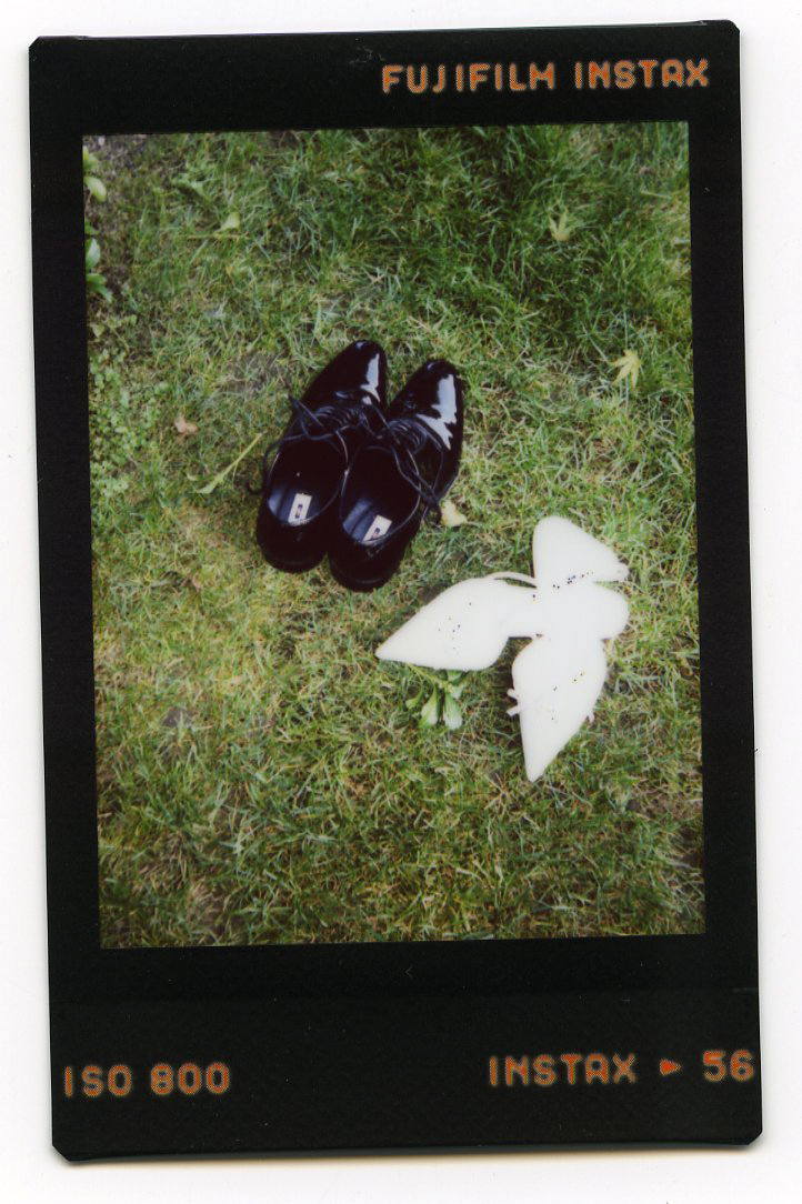A pair of shiny black dress shoes and a white origami dove rest on green grass in a Fujifilm Instax instant photo, capturing the charm often seen in Glen Manor House Wedding Photography.