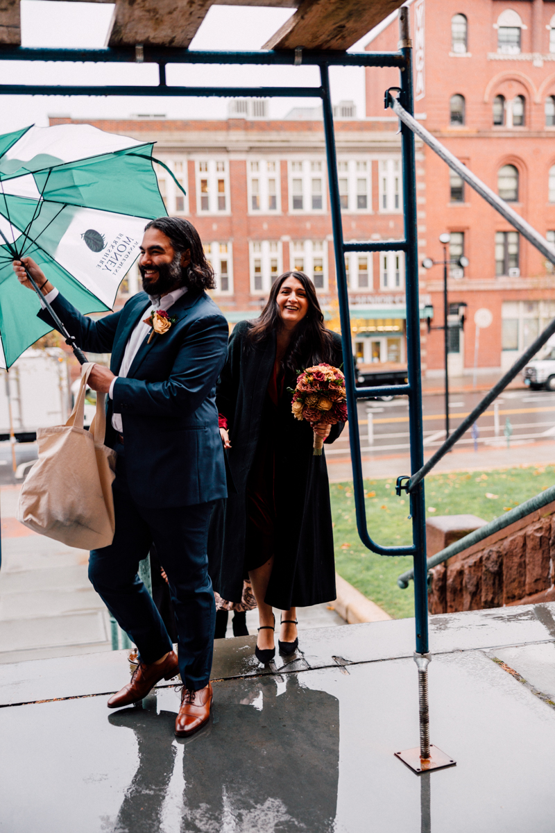 A man holds a green and white umbrella while walking with a woman in a formal outfit, both smiling as they approach a building during a rainy day.