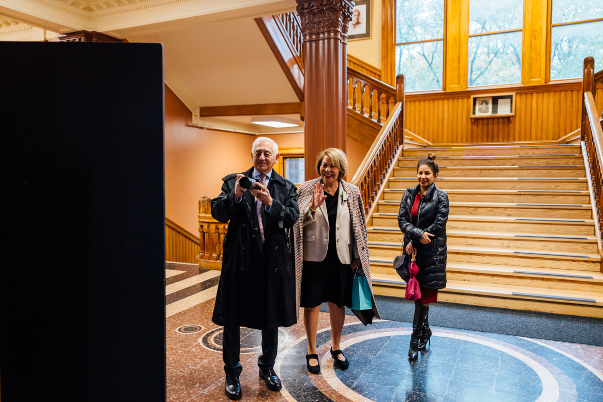 Three individuals interacting in a historic building with wooden stairs and large windows, capturing a moment together.