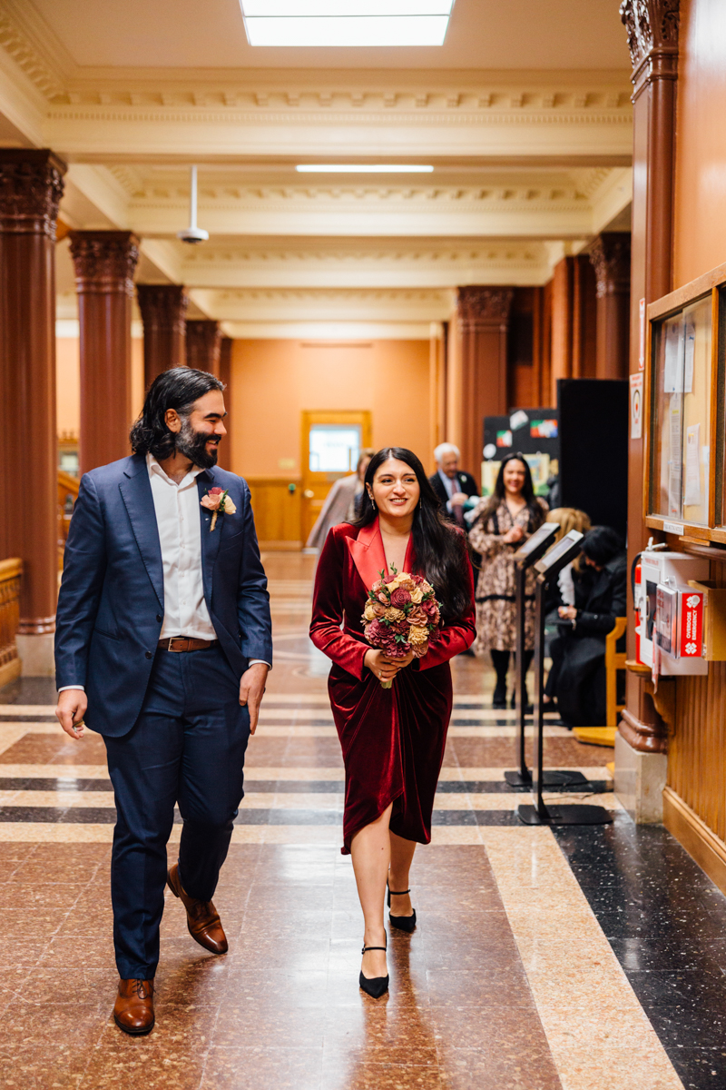 A couple strolls through a beautifully decorated hallway, the woman in a red velvet dress holding a bouquet, while the man in a suit smiles beside her.