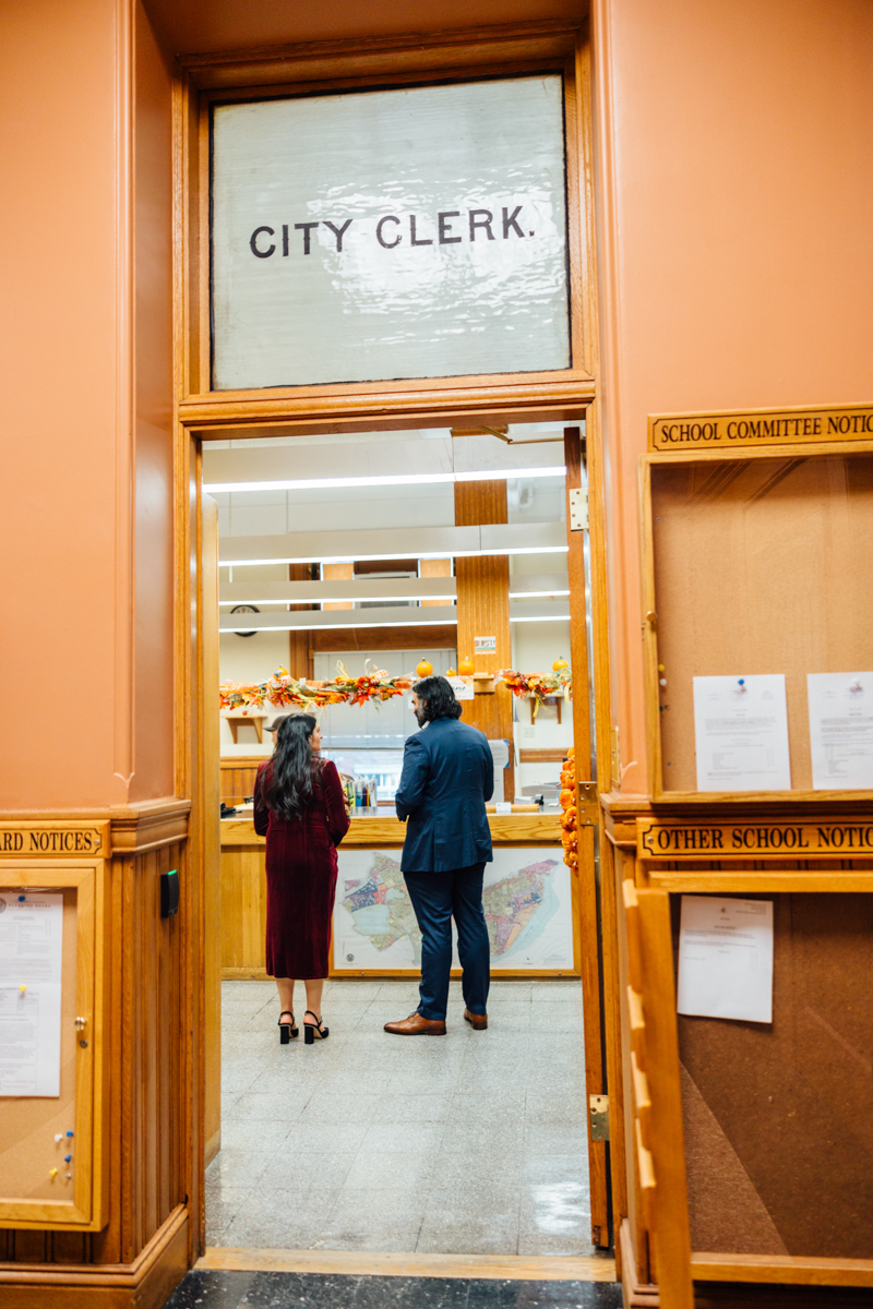 Two individuals converse at the counter of the City Clerk's office, with various notices displayed nearby.