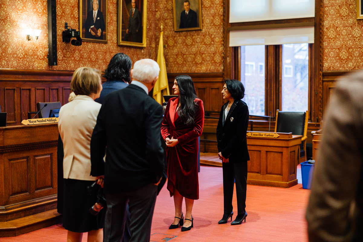 A group of individuals in formal attire conversing inside a historic city hall, featuring portraits on the walls and an official setting.