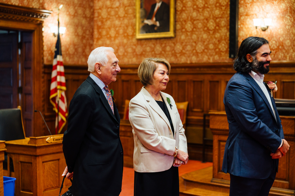 Three individuals stand attentively in a beautifully decorated room with ornate wallpaper, an American flag in the background, and a portrait on the wall.