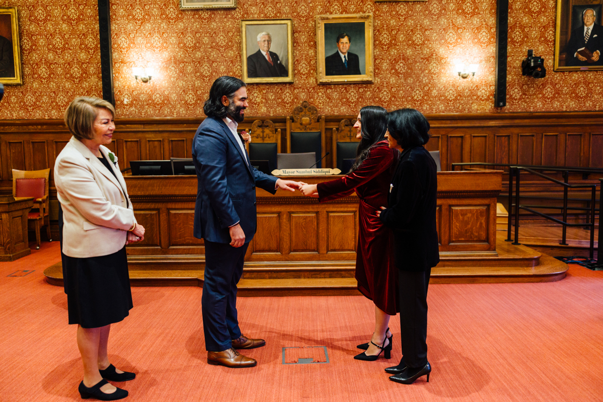A couple exchanges vows in a formal setting, surrounded by family and officials, with portraits hanging on the wall.