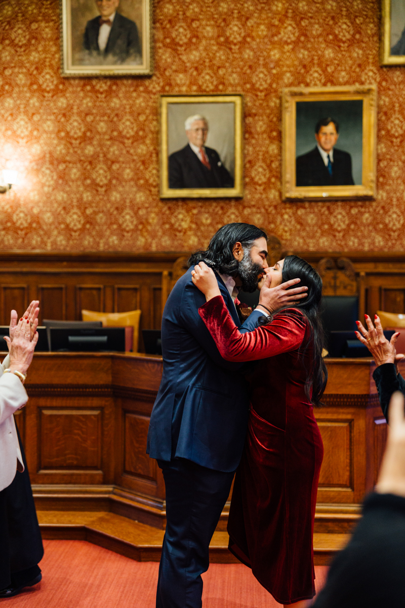 A couple shares a kiss in a historic room adorned with portraits, surrounded by supportive guests.