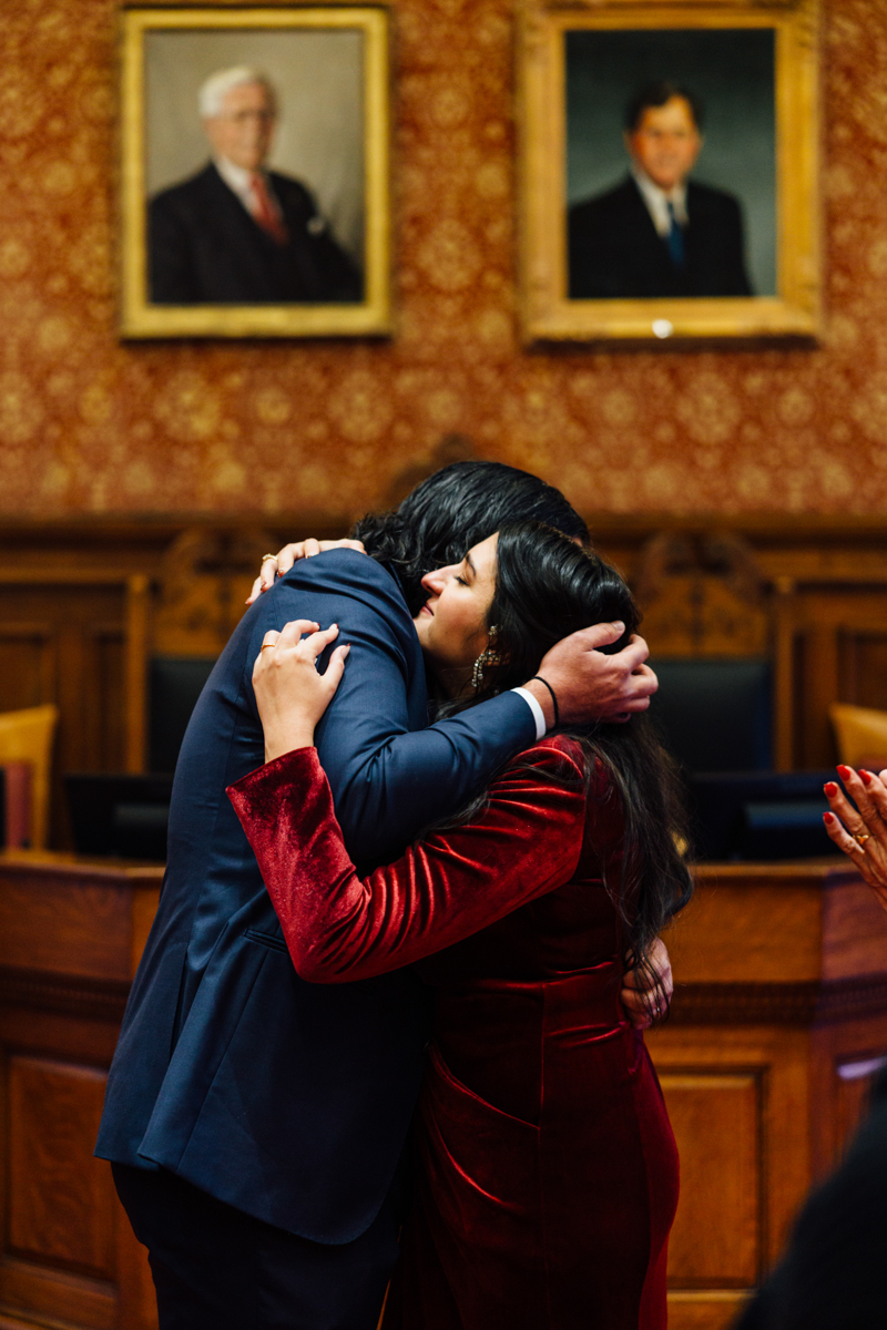 A heartfelt embrace between two individuals in formal attire within a historic room adorned with portraits, capturing a moment of joy and celebration.