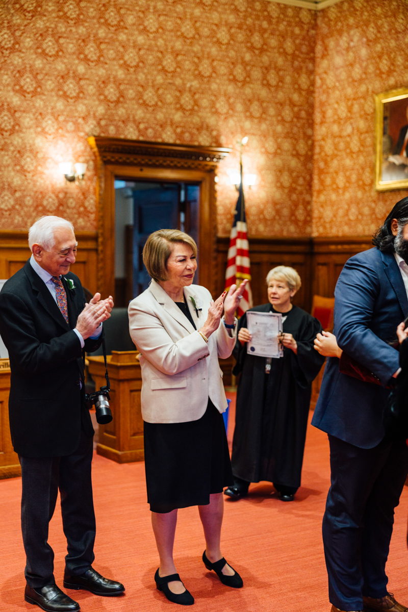 A group of individuals in formal attire engaging in a celebratory moment in a courtroom, with an official in a robe holding documents in the background.