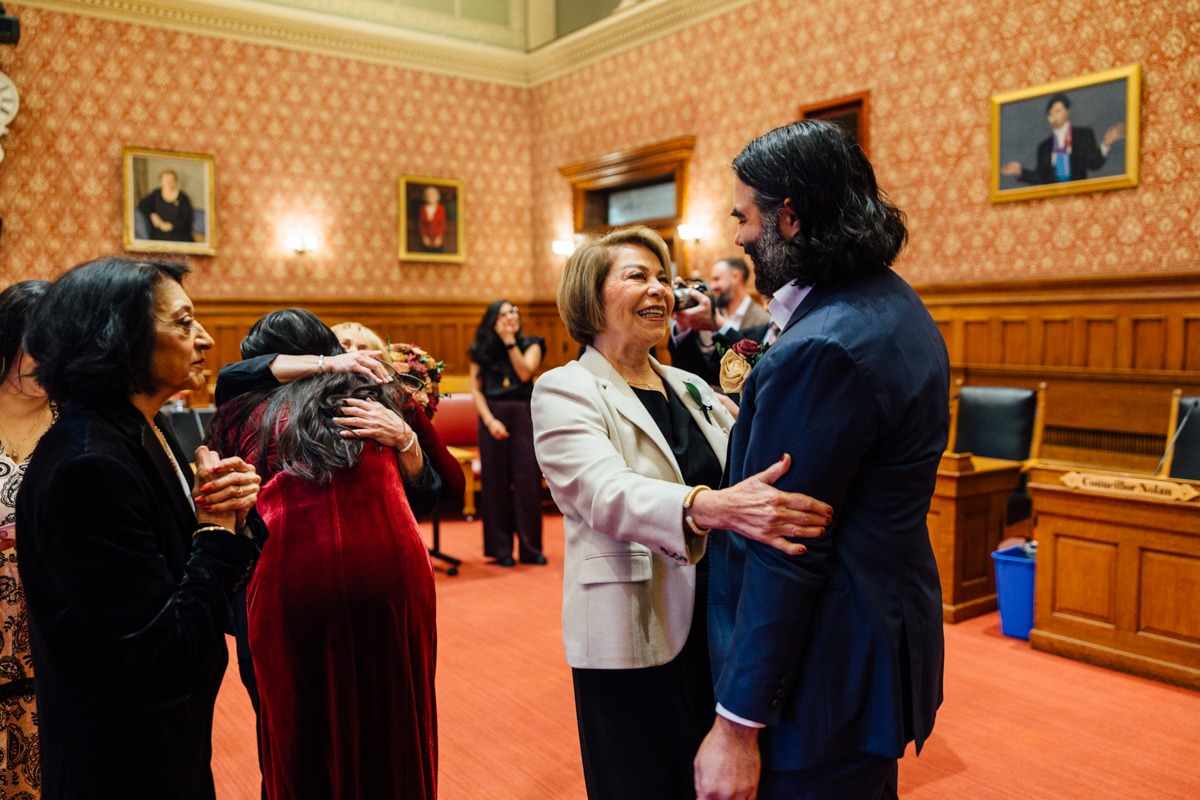 A joyful interaction between a man and a woman in formal attire, surrounded by family and friends, capturing a heartfelt celebration in an elegant room adorned with historical portraits.