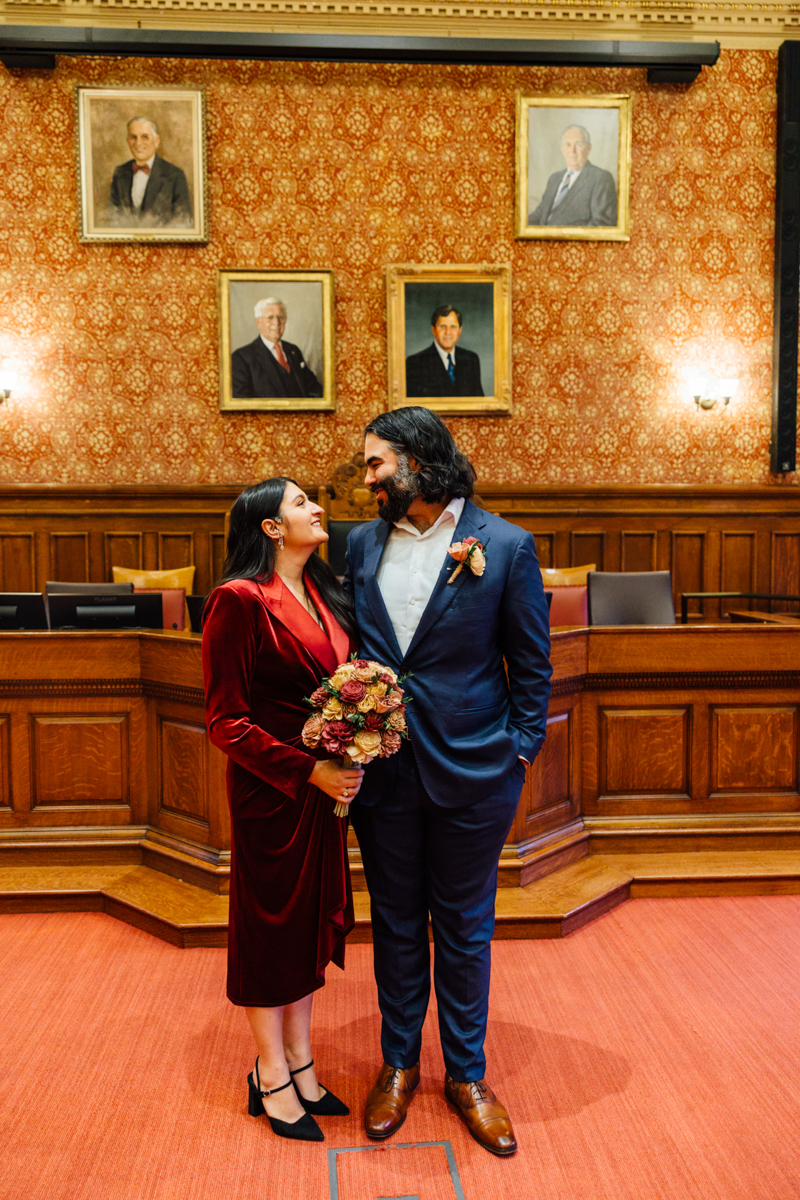 A joyful couple stands close together, smiling at each other in a beautifully decorated historic room, adorned with portraits and an elegant ambiance, as they celebrate their love.