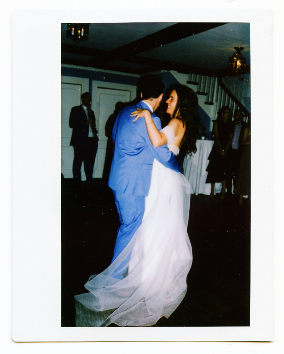 A bride in a flowing white dress and a groom in a blue suit dance together, smiling and gazing at each other, during their Dennis Inn Wedding in Dennis, MA, with guests and a staircase visible in the background.