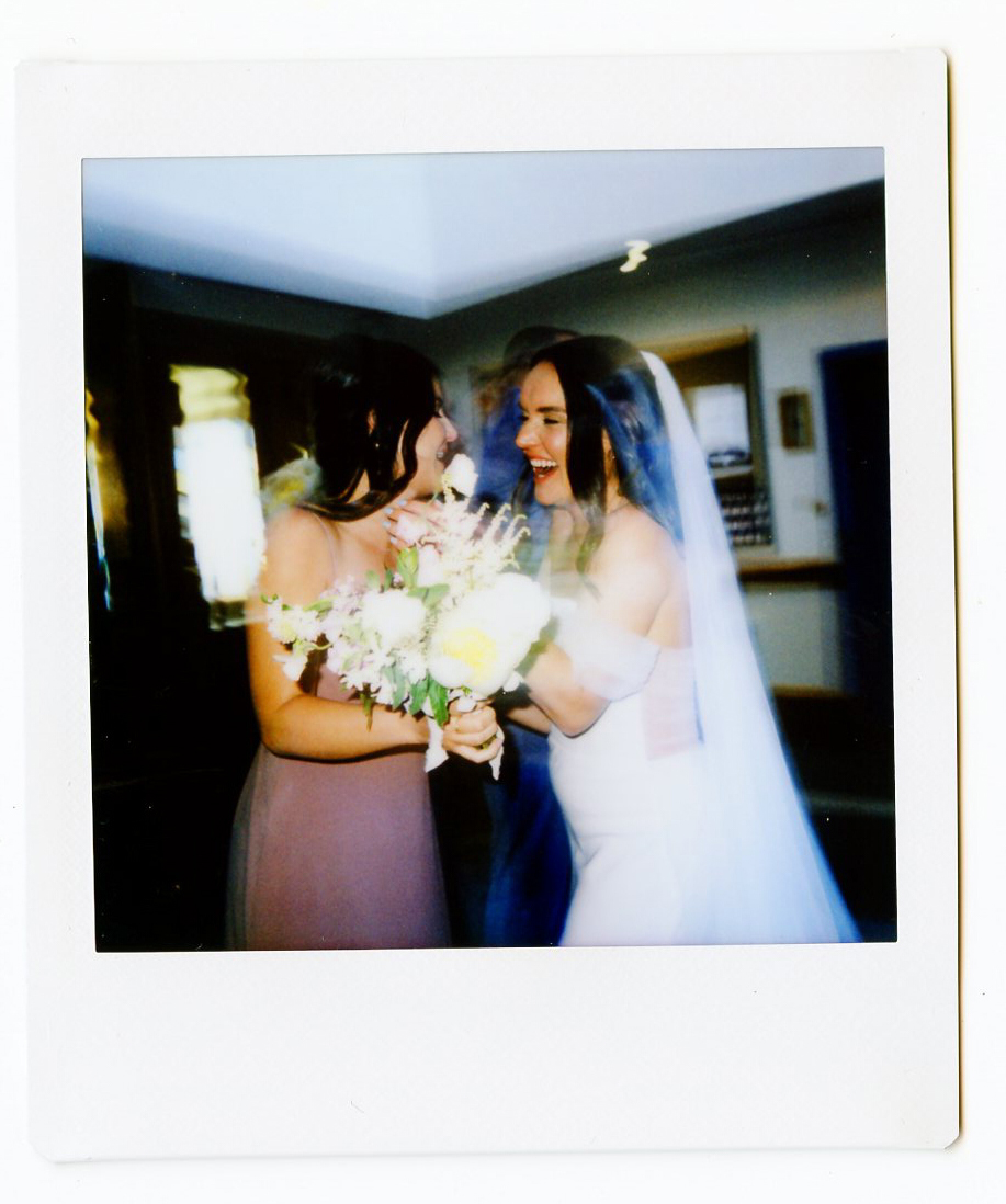 A bride in a white dress and veil smiles joyfully, holding hands with a bridesmaid in mauve at their Dennis Inn Wedding, the excitement captured indoors with blurred motion.