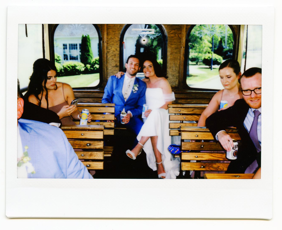 A bride and groom sit together smiling on a wooden trolley at their Dennis Inn Wedding, friends around them. The bride wears a white dress; the groom is in a blue suit. Green Dennis MA scenery shows through the windows.