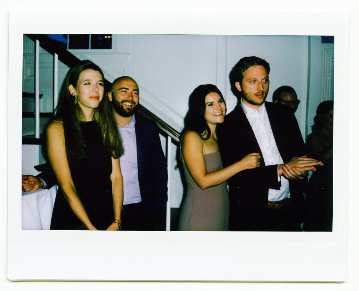 Four people dressed in formal attire stand closely together indoors, smiling and looking to the right. A staircase and white walls are in the background, suggesting a joyful Cape Cod Wedding or celebration at the Dennis Inn Wedding venue.