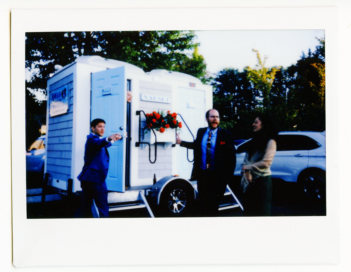 Three people dressed formally stand in front of a white restroom trailer decorated with a red ribbon and bow at a Dennis Inn Wedding in Dennis, MA. One man points outward, while another holds giant scissors for the ribbon-cutting ceremony.