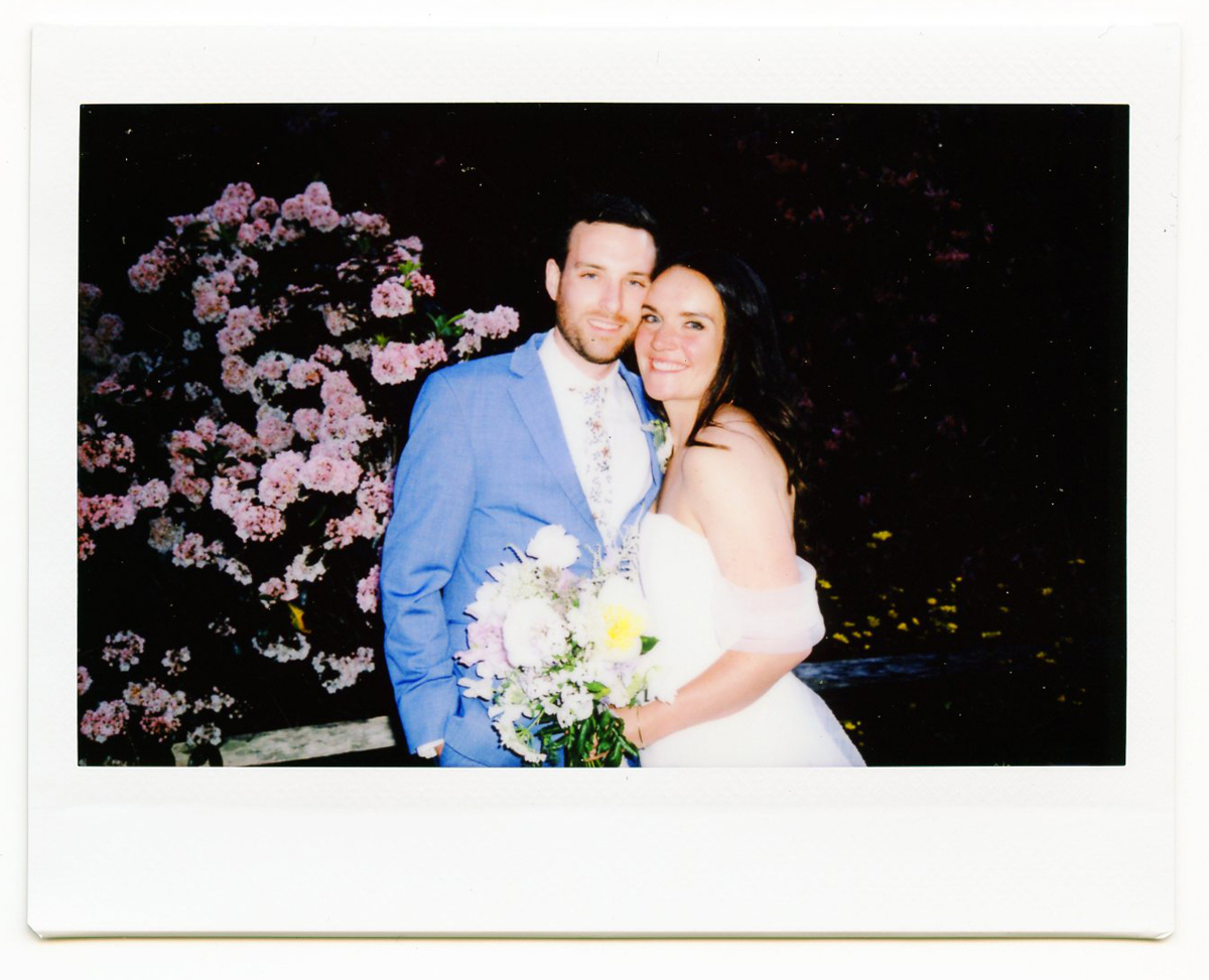 A smiling couple in wedding attire stands close together outdoors at their Dennis Inn Wedding; the groom sports a blue suit and floral tie, while the bride in a white off-shoulder dress holds a bouquet. Pink flowers bloom in the background.