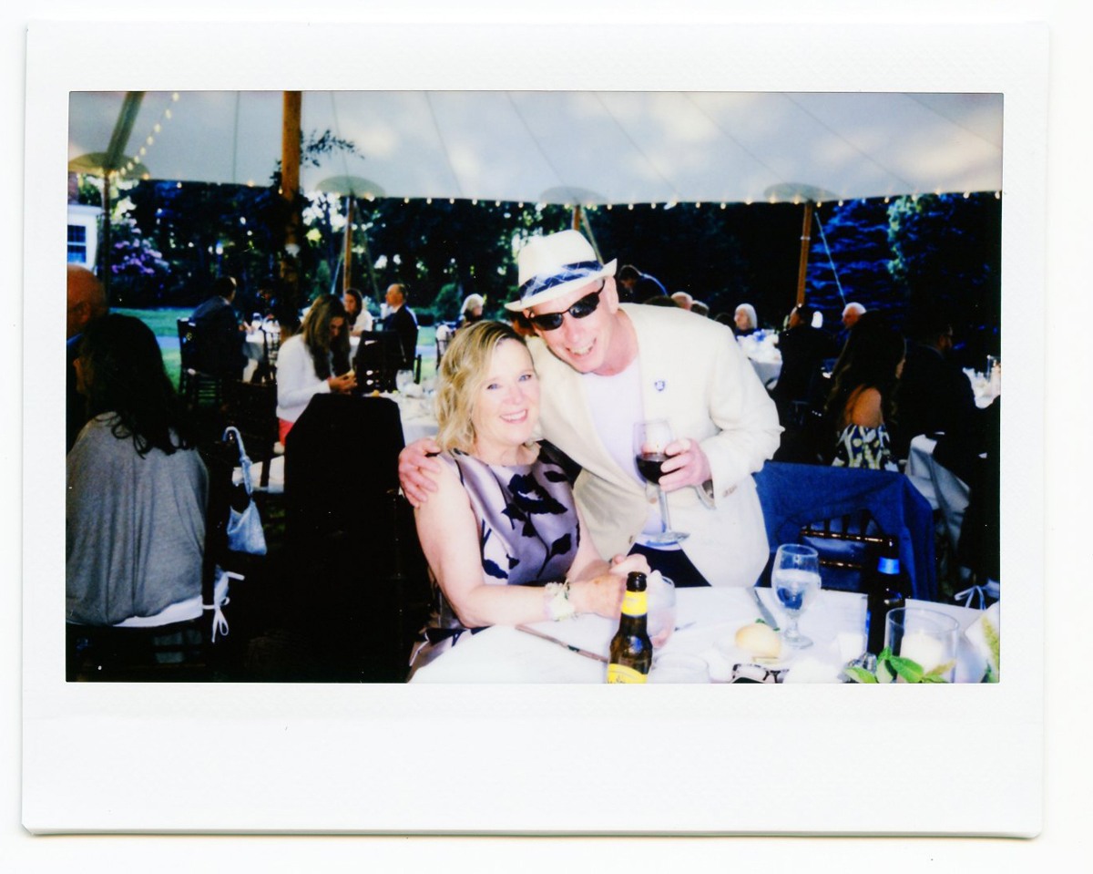 A man in a light suit and hat hugs a smiling woman seated at a table with drinks at an outdoor Cape Cod wedding under a tent, with other guests visible in the background.