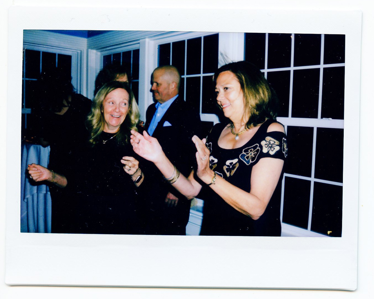 Three adults, two women and one man, are dressed formally and appear to be dancing or clapping at a lively Dennis Inn wedding in Dennis, MA. Large windows in the background add to the cheerful indoor atmosphere.