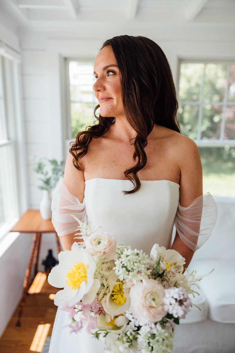 A bride with long brown hair in loose waves, wearing an off-the-shoulder white gown, holds a bouquet of white and yellow flowers while standing indoors and looking out a sunlit window.