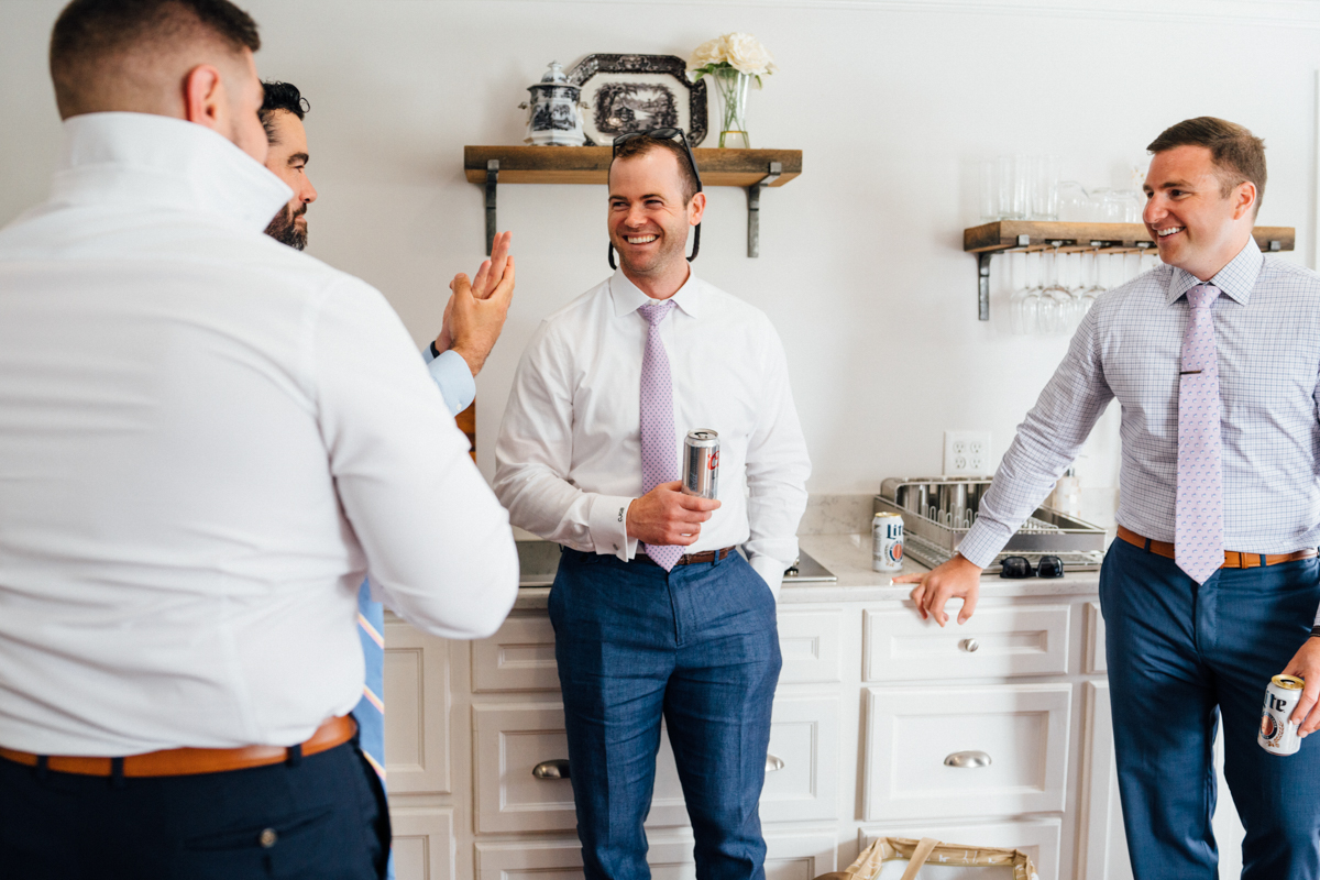 Four men in dress shirts and ties stand in a bright kitchen, smiling and talking. Three hold cans of beer, and one adjusts his collar. Shelves with decor and glasses are in the background.