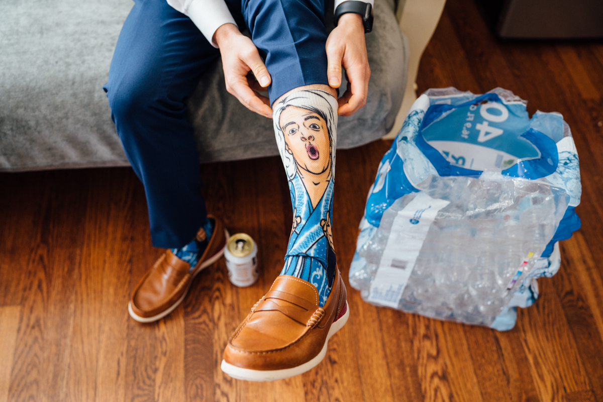 A person in brown loafers and blue pants lifts a pant leg to reveal a colorful sock featuring a surprised cartoon woman. A can and a pack of bottled water are on the wooden floor nearby.