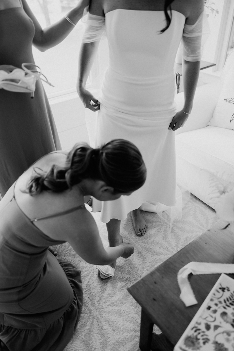 A bride in an off-the-shoulder dress stands as one woman kneels to help with her shoe and another stands nearby holding another shoe. The scene is indoors, filled with soft natural light.