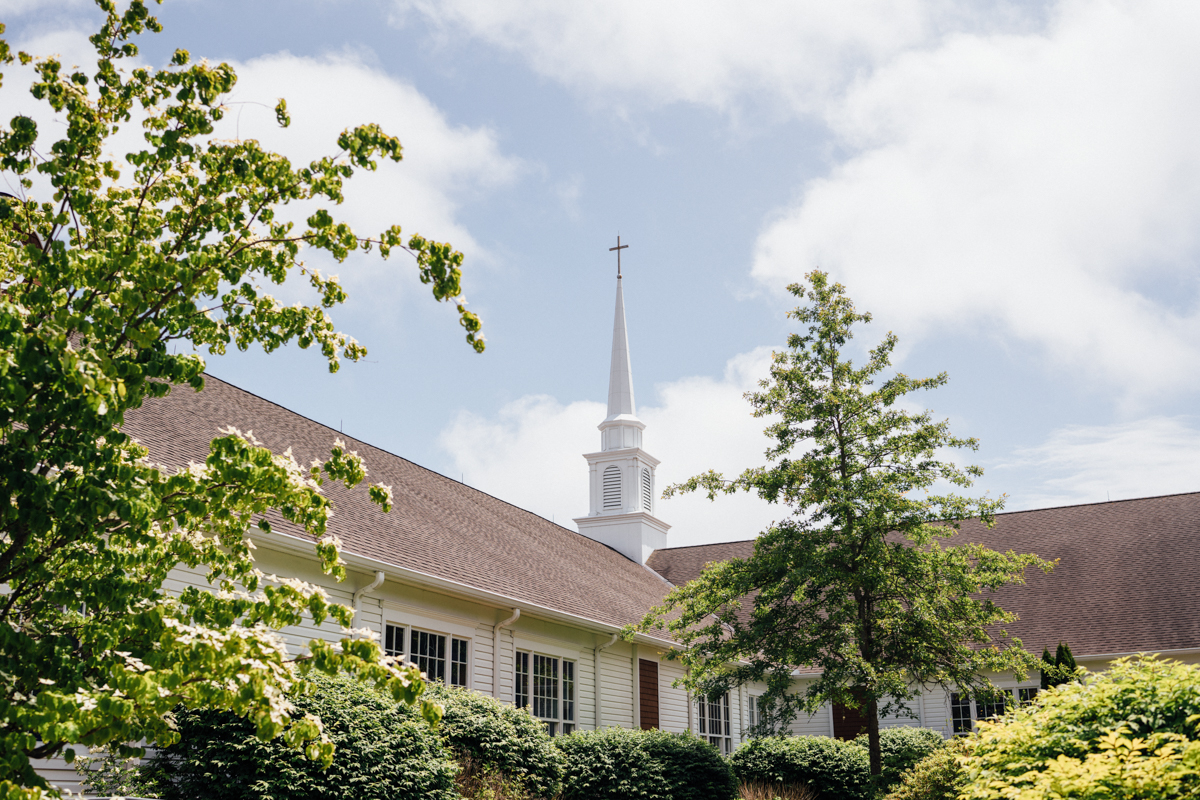 A white church building with a tall steeple and cross, surrounded by green trees and shrubs under a partly cloudy sky.