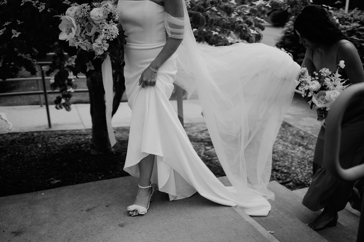 A bride in a long, flowing wedding gown is walking outdoors, holding up her dress and bouquet. Another woman follows behind, holding the train and carrying flowers. The image is in black and white.