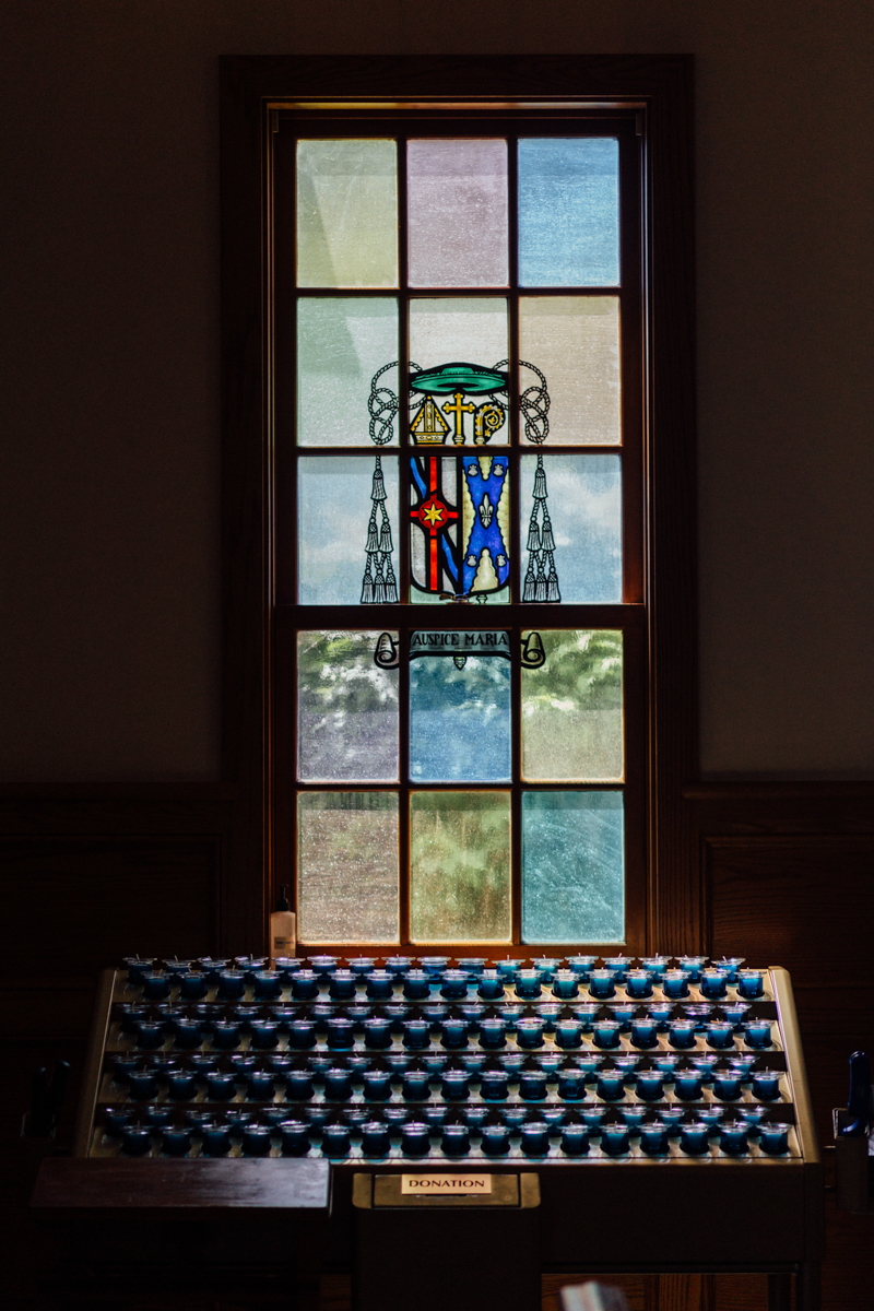 A stained glass window with religious symbols and the words AMARE ET SERVIRE above a row of unlit prayer candles in a dimly lit room.