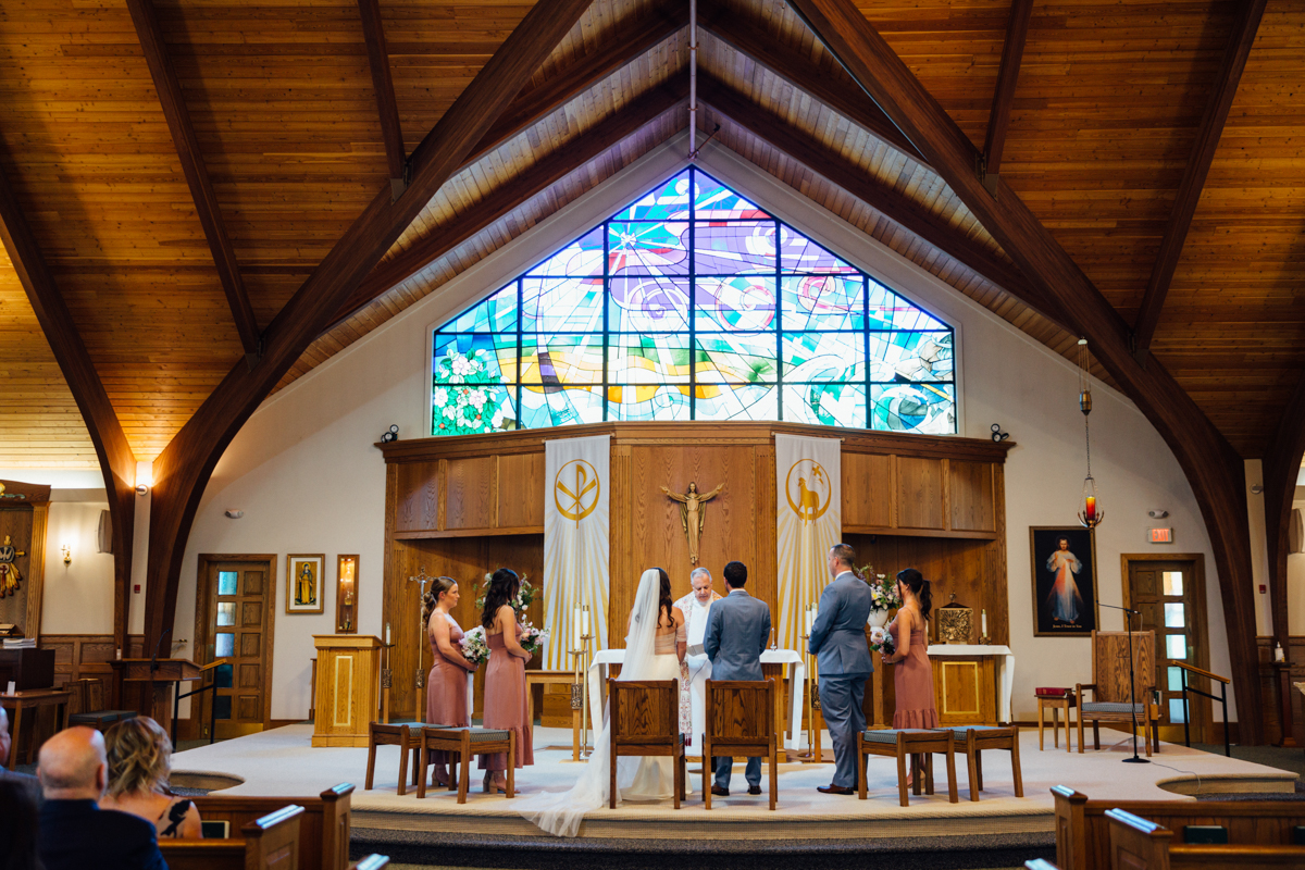A wedding ceremony takes place in a church with a wooden ceiling and colorful stained glass window. The bride and groom stand at the altar with an officiant as bridesmaids and groomsmen look on. Guests are seated in pews.