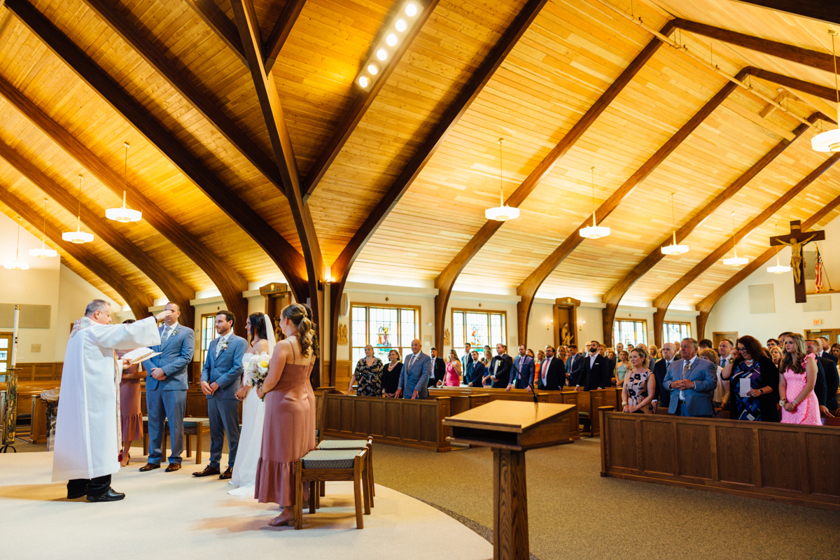A wedding ceremony inside a church. A priest stands before a bride, groom, and wedding party, while guests in formal attire watch from pews. Sunlight streams in through stained glass windows under a high wooden ceiling.