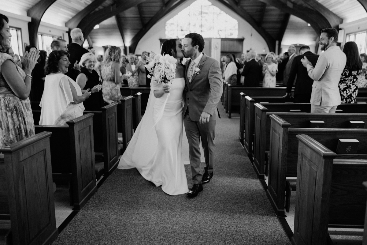 A bride and groom walk down the aisle of a church, surrounded by guests who are standing, clapping, and smiling. The scene is joyful, and the couple shares a kiss as they exit the ceremony.