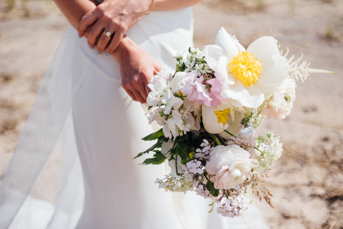A bride in a white dress holds a bouquet of white and pale pink flowers with greenery, standing outdoors on a sunny Dennis Inn Wedding day. Only her torso and arms are visible.