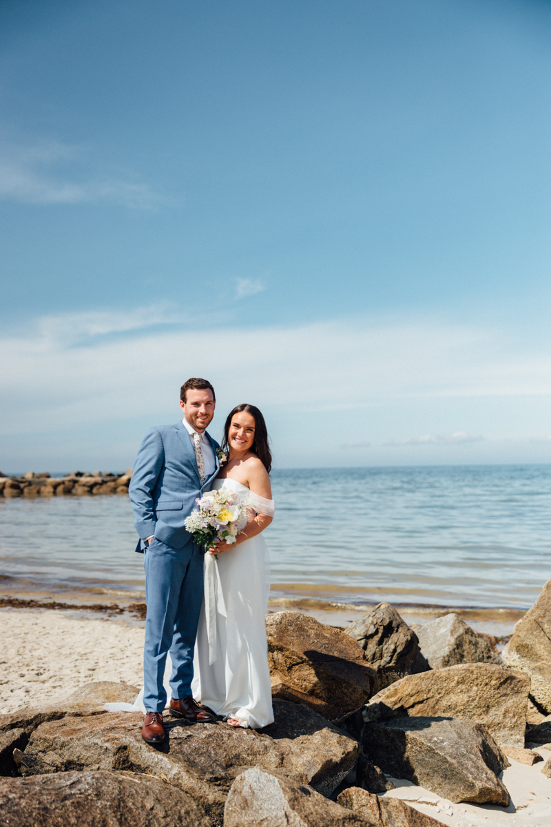 A bride in a white dress and a groom in a blue suit stand on rocks by the ocean in Dennis MA, smiling and holding a bouquet, with blue sky and calm water in the background—capturing the joy of their Dennis Inn Wedding.