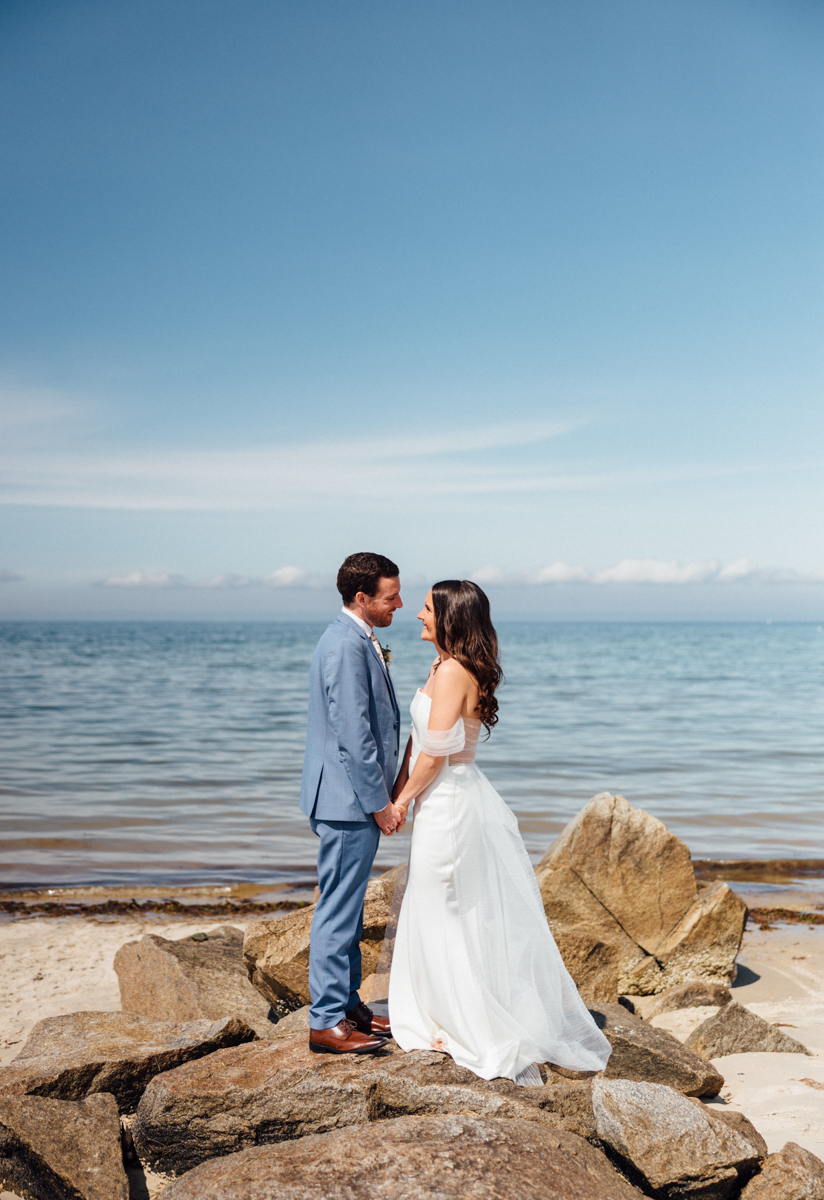 A bride and groom stand on rocks at a Dennis Inn Wedding in Dennis, MA, holding hands and facing each other with calm water and a blue sky behind them. The bride wears a white gown, and the groom wears a light blue suit.