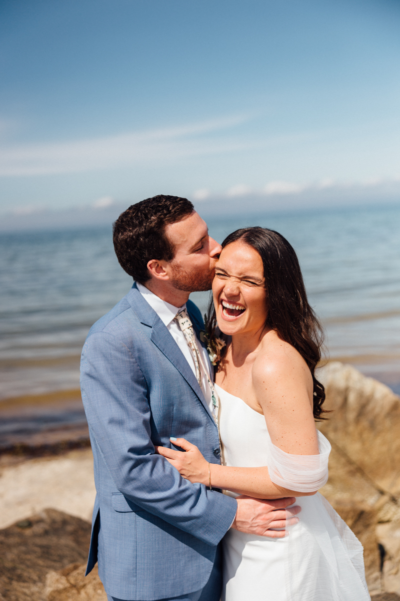 A groom in a light blue suit kisses his laughing bride on the forehead. They stand close together, smiling, at their Dennis Inn wedding in Dennis, MA, with a calm ocean and clear blue sky as their backdrop.