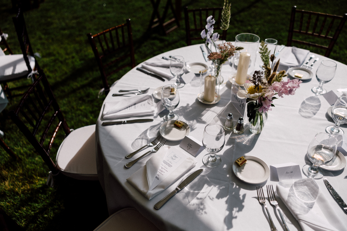 A round table set outdoors at a Dennis Inn Wedding features a white tablecloth, neatly arranged plates, cutlery, glasses, napkins, menus, candles, and small floral centerpieces. Sunlight casts soft shadows across the table and chairs.
