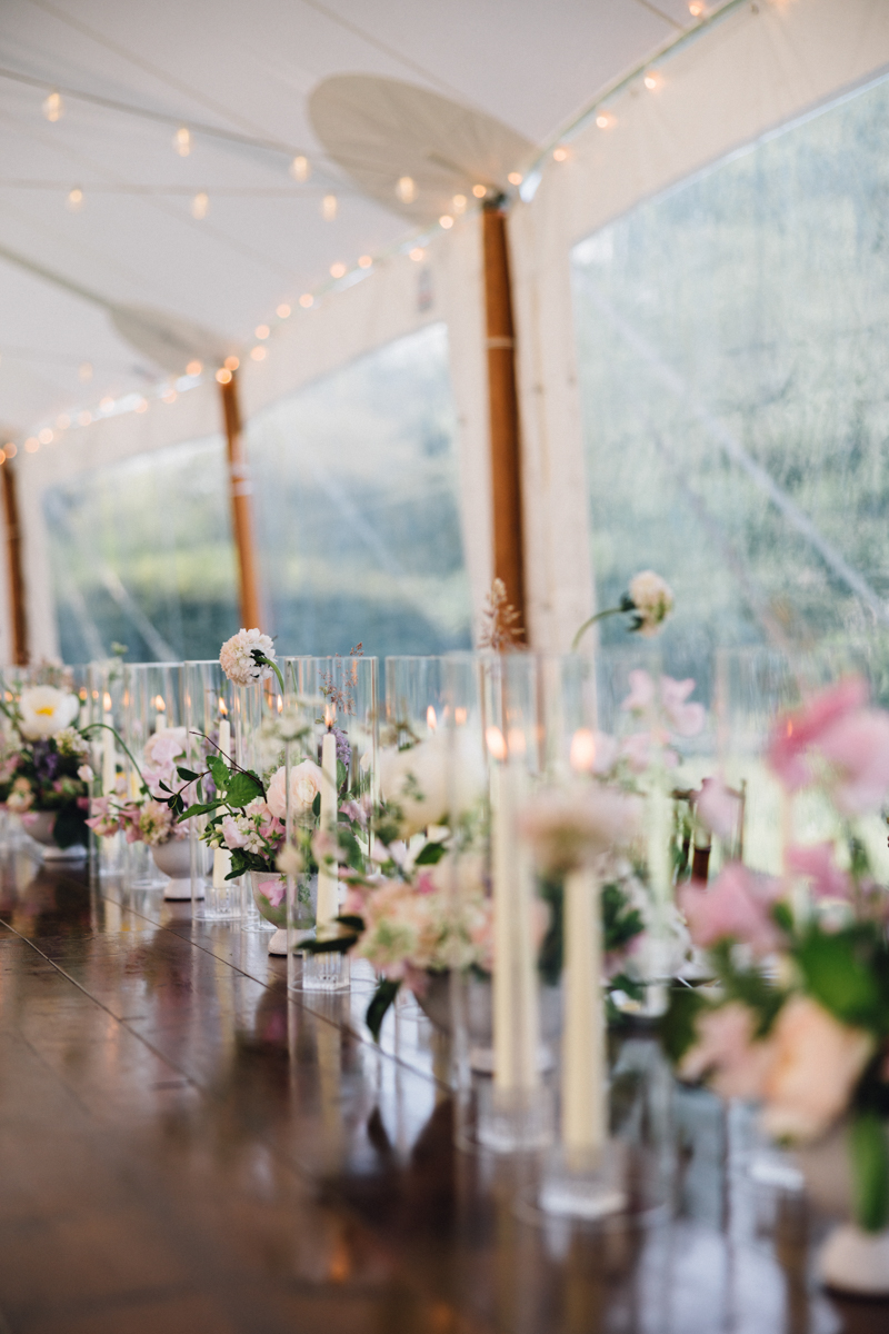 A long wooden table at a Dennis Inn Wedding is decorated with glass vases of pink and white flowers, tall white candles, and set under a white tent with soft string lights and large windows revealing lush greenery outside.