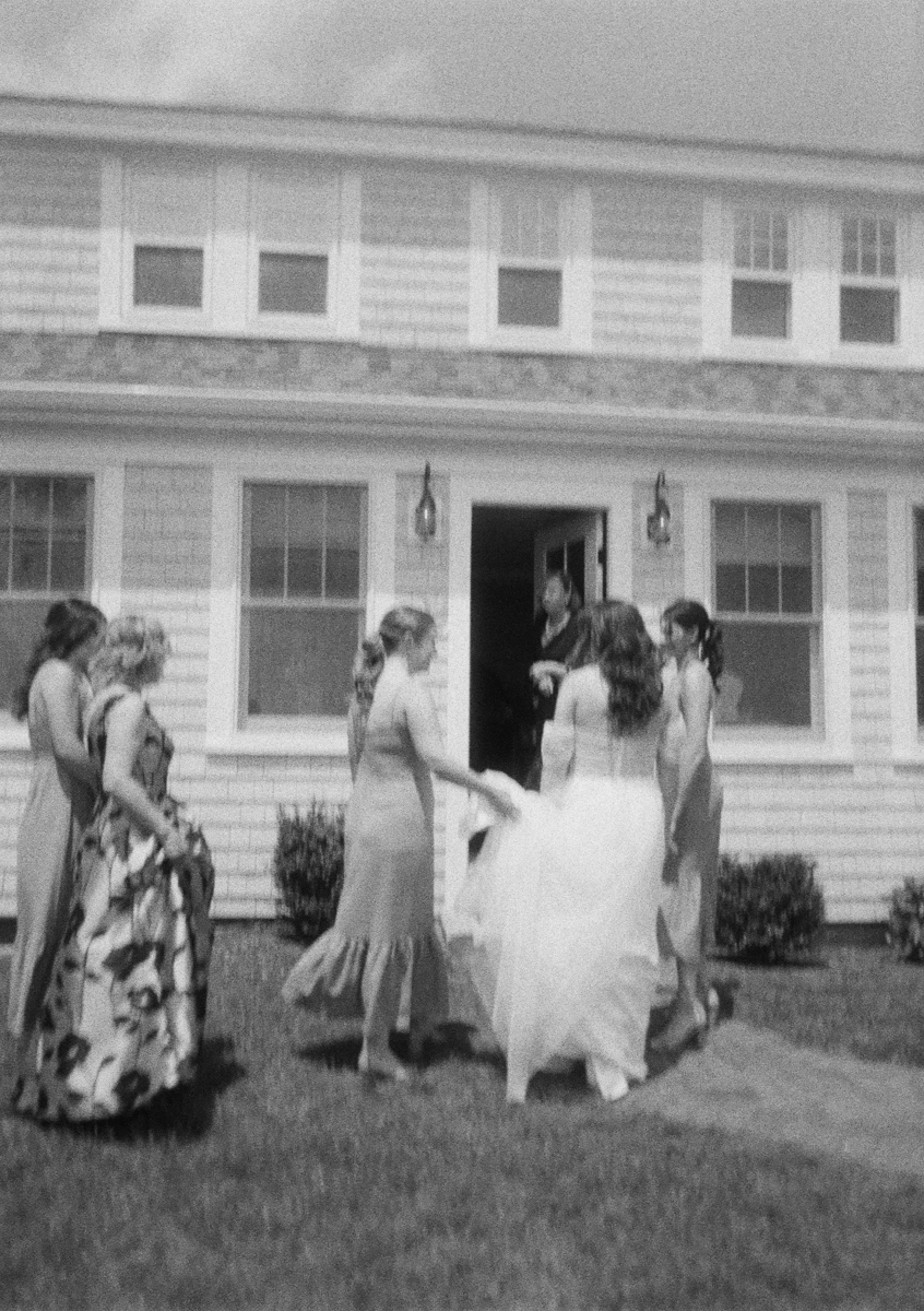 A black-and-white photo of a bride in a flowing dress with her bridesmaids, outside a house at what could be a Dennis Inn Wedding. The group faces the open front door, where someone inside appears to greet them.