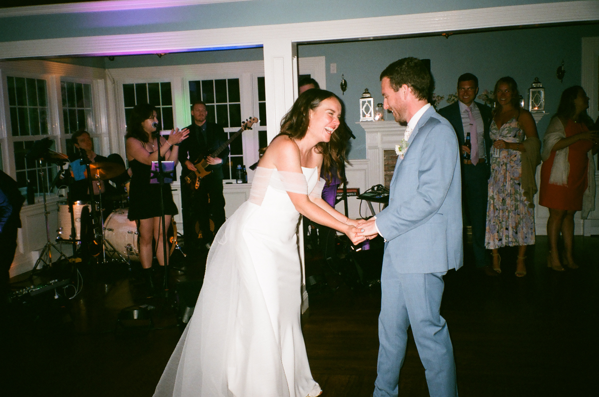 A bride in a white dress and a groom in a light blue suit laugh and hold hands while dancing at their Dennis Inn Wedding reception, with guests and a live band adding to the joyful atmosphere in the background.