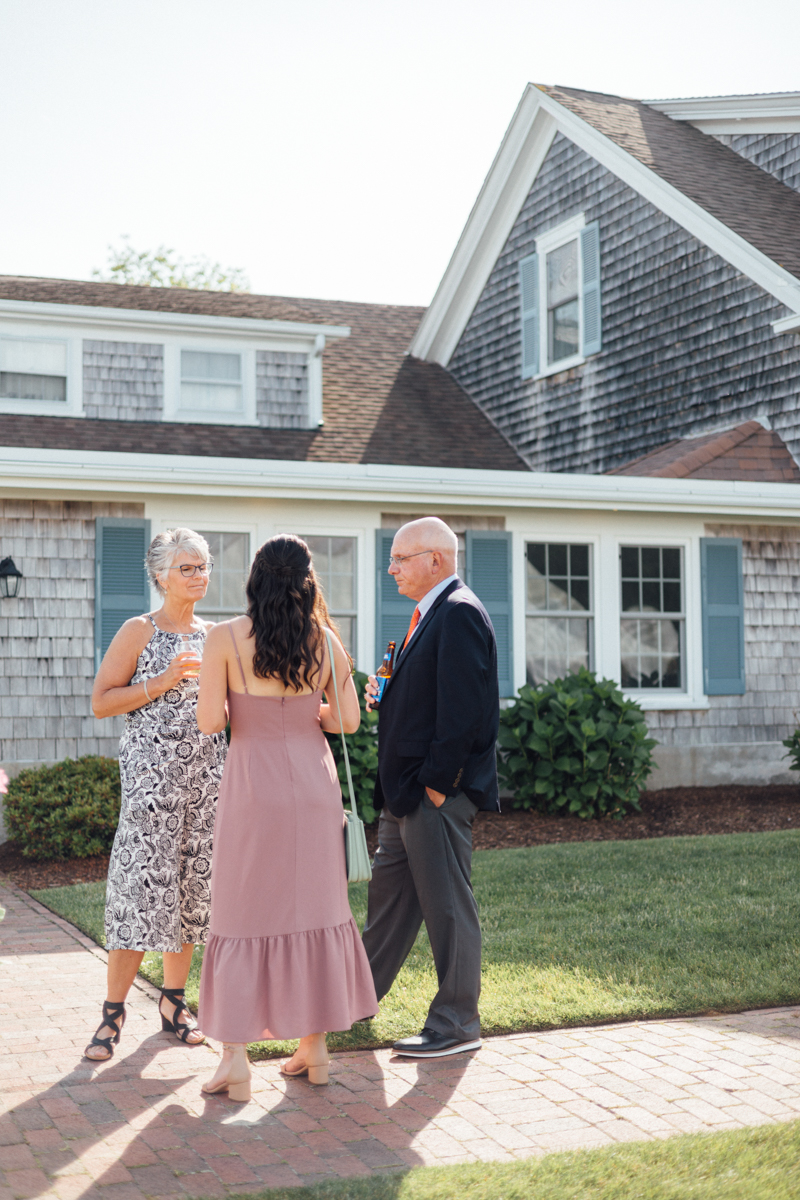 Three adults stand outside a shingle-style house, engaged in conversation at a Dennis Inn Wedding. One woman wears a patterned dress, another a mauve dress, and the man is in a suit and tie. The sunny setting feels relaxed.