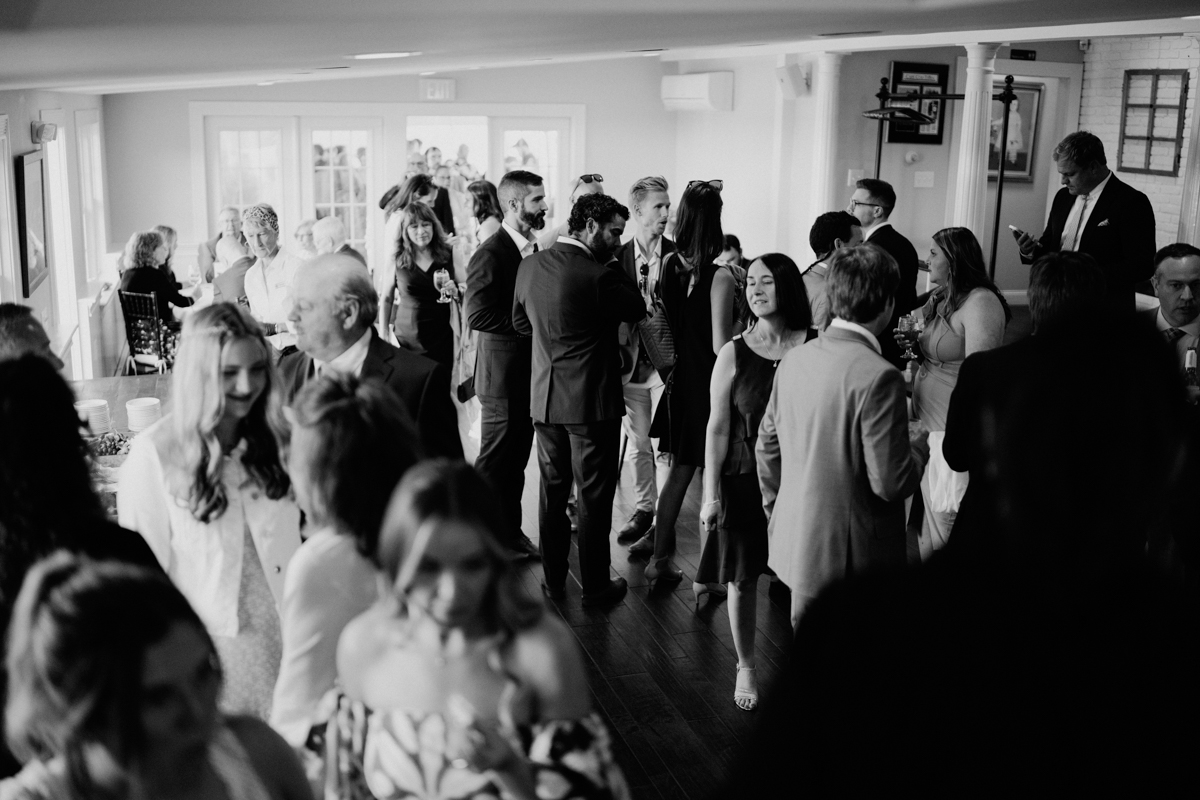 Black and white photo of a lively Dennis Inn Wedding, with guests dressed formally, mingling and chatting in a well-lit room with large windows in the background. Some guests are holding drinks.