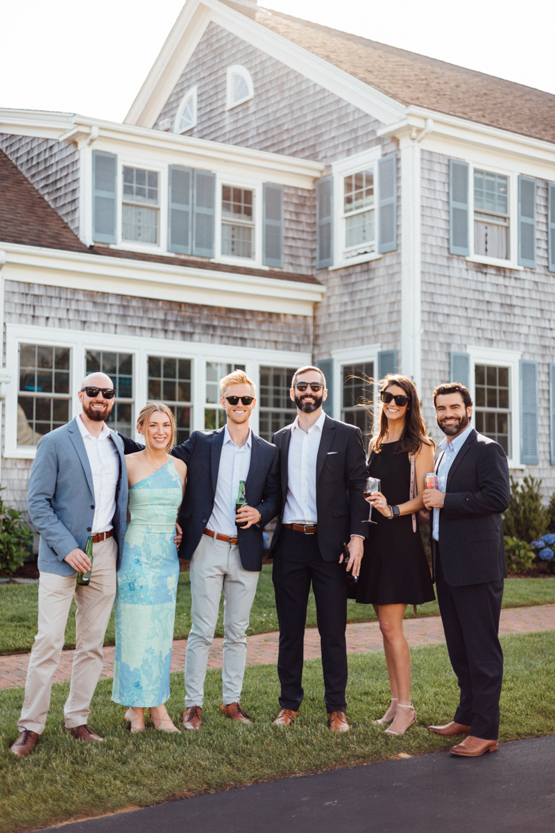Six adults dressed in semi-formal attire stand smiling with drinks in hand, posing for a photo on a sunny day in front of a large shingle-style house with blue shutters after a beautiful Dennis Inn Wedding.