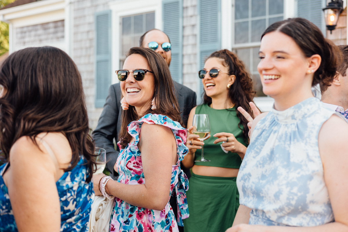 A group of people outdoors, smiling and laughing at a Dennis Inn Wedding. One woman in sunglasses wears a floral dress, another in green holds a wine glass, and others wear patterned outfits. The festive scene unfolds with a house in the background.