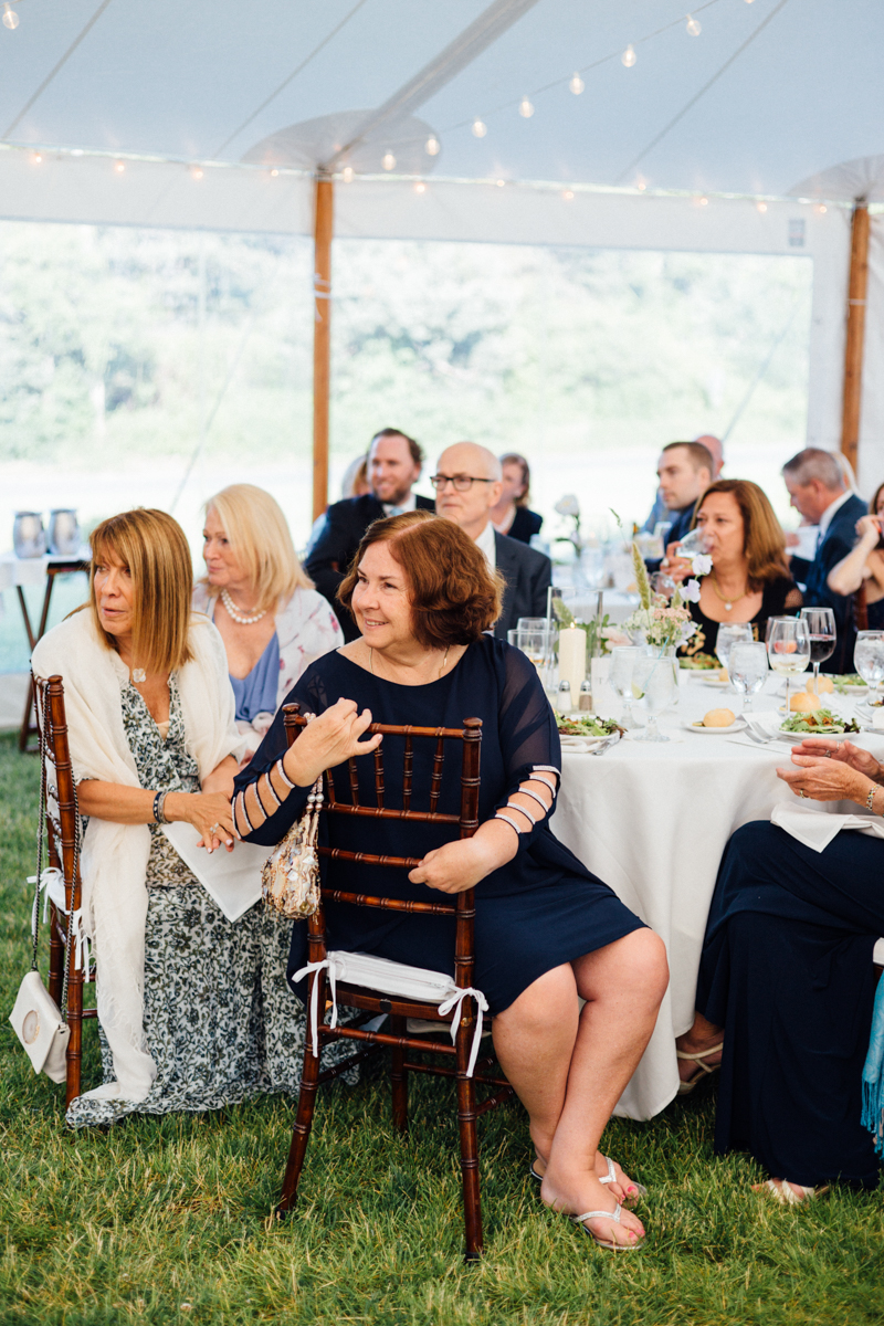 Guests in formal attire sit at round tables under a white tent at a Dennis Inn Wedding, likely a reception. They appear engaged and are surrounded by elegant table settings, flowers, and string lights overhead.