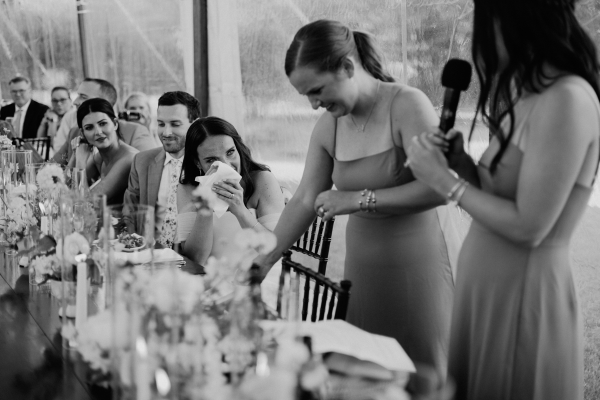 A woman wipes her eyes with a tissue, appearing emotional, as another woman speaks into a microphone at a long, decorated table during what seems to be a Dennis Inn Wedding on Cape Cod, with guests seated and watching the heartfelt moment unfold.