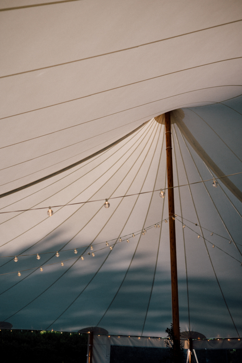 A close-up view of the inside of a large white event tent at a Dennis Inn Wedding in Dennis, MA, featuring a central wooden pole and string lights hanging across the ceiling.